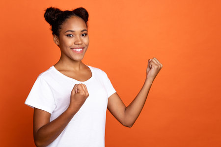 Photo of young lovely african woman rejoice victory triumph awesome fists hands isolated over orange color backgroundの写真素材