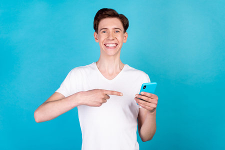 Photo of young guy indicate finger smartphone advertise recommend suggest isolated over blue color...