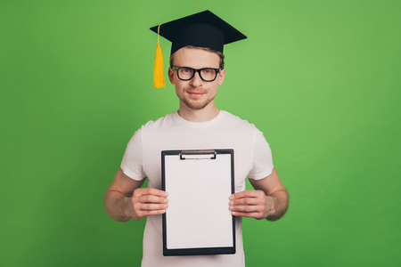Photo of a-student man hold clipboard fill exchange form wear mortar board cap glasses on green color backgroundの写真素材