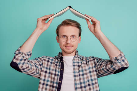 Funny nerd guy hold book open above head look camera isolated on teal backgroundの写真素材