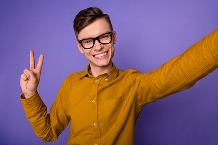 Photo of youth excited man shooting selfie show fingers peace cool v-symbol isolated over violet color backgroundの写真素材