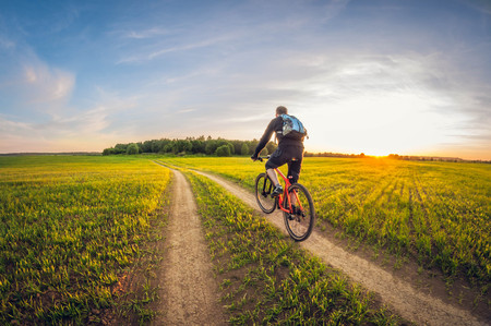 Cyclist on a dirt road in a field at sunset, cycling on a summer evening, sown field landscapeの写真素材