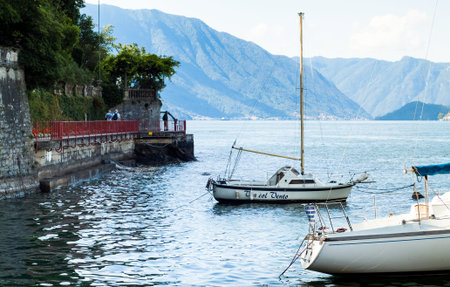 Varenna, Italy - September 4th, 2015: tourists walk along an escapade in Varenna (Lake Como, Italy), with two white boats in the foreground.のeditorial素材
