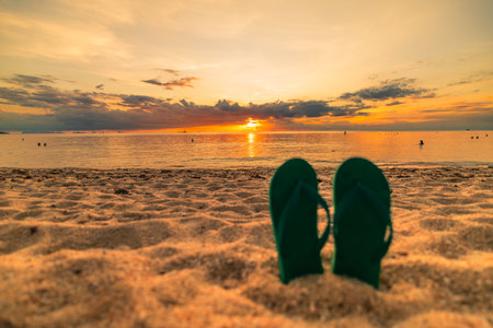An image featuring green flip-flops resting on the sand as the sun sets, reflecting the relaxed and beautiful atmosphere of summer at the beach. In the background, people are enjoying a special moment by the sea, further enhancing this picture with good memories of a holiday.の写真素材