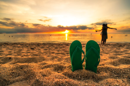 An image featuring green flip-flops resting on the sand as the sun sets, reflecting the relaxed and beautiful atmosphere of summer at the beach. In the background, people are enjoying a special moment by the sea, further enhancing this picture with good memories of a holiday.の写真素材