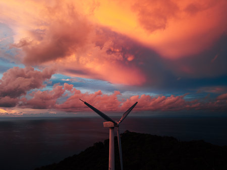 wind turbine prominently situated on a hilltop, silhouetted against a dramatic sunset sky filled with thick orange-red and blue cloudsの写真素材