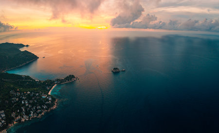aerial shot of Koh Tao, Thailand, at dawn. The morning sun casts a golden glow upon the calm sea, contrasting with the soft orange and pink hues of the skyの写真素材