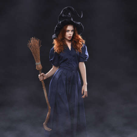 Young beautiful red-haired witch with a broom in the dark fog
