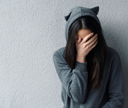 Sad woman in grey hoodie with cat's ears covering face with hand. Wall background.