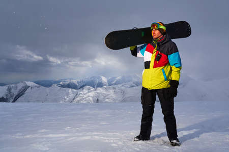 Snowboarder carries a snowboard in hand. Snowfall on the background of mountains, ski resort.の写真素材
