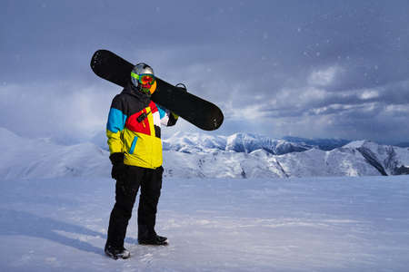 Snowboarder carries a snowboard in hand. Snowfall on the background of mountains, ski resort.の写真素材