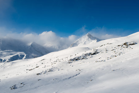 landscape of a gentle slope with rocks against a background of snow-capped mountains with clouds