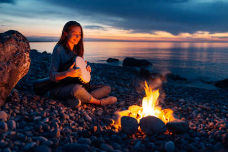 cheerful percussionist girl playing djembe sitting on the beach by the fire at sunset.の写真素材