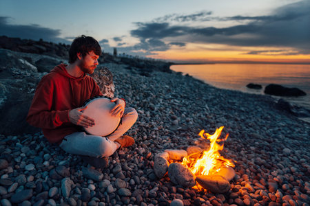 percussionist guy plays a djembe sitting on the beach by the fire at sunset.