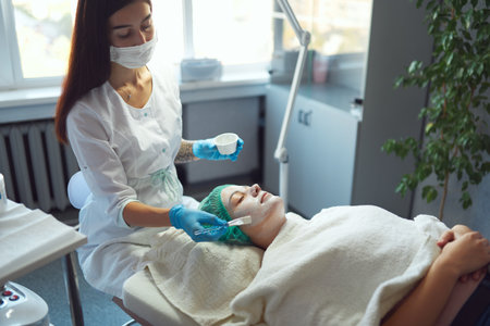 Beautician covering woman facial skin with moisturizing cleansing mask during skin care procedure in a beauty salon.の写真素材
