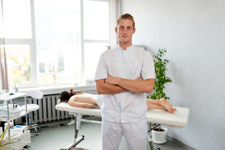The masseur stands with crossed arms in the medical center, the girl behind him lies on the couch. Healing massage and body care.の写真素材
