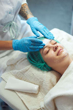 Beautician pleasantly massages with rub peeling cream face of woman on couch. Deep cleansing facial.の写真素材