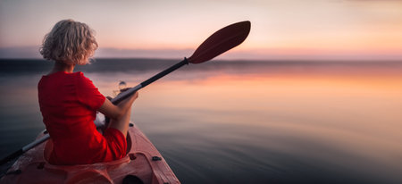 Blonde woman in a red dress sits in a boat with a paddle at sunset.の写真素材