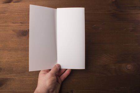 Blank tri fold brochure in hand on wooden background to replace your design or message. A mock-up for brand identification for designersの写真素材