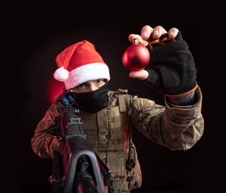 Concept shoot about the man holding Christmas ball with machine gun and wearing Christmas hat on...