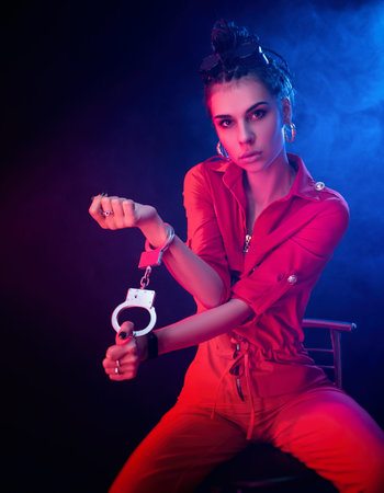 a bright brunette girl in a red jumpsuit handcuffed on a dark background in neon light
