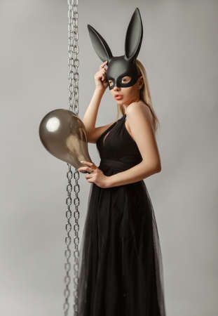 Beautiful blonde in a rabbit mask in a sexy evening dress on a white background with a metal chainの写真素材