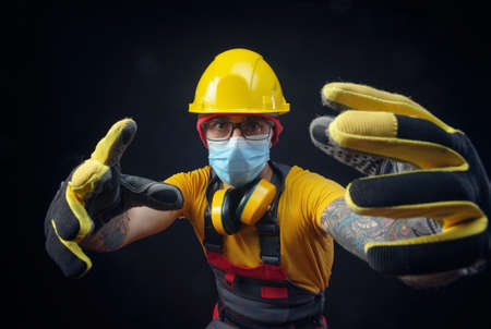 a man in a construction repairmans overalls wearing a medical mask against the flu coronavirus and a hard hat on his headの写真素材