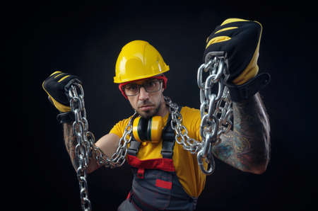 a man in the overalls of a construction repairman with a medical mask against coronavirus holds a metal chain in his handsの写真素材