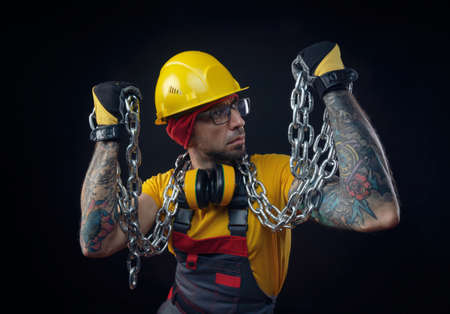 a man in the overalls of a construction repairman with a medical mask against coronavirus holds a metal chain in his handsの写真素材