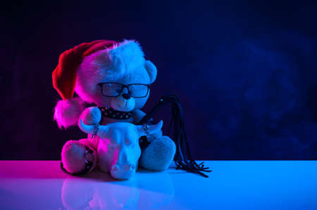 a toy bear in a Santa Claus hat wearing leather straps harness accessory for BDSM along with a statuette of a white bull on a dark background in neon lightの写真素材
