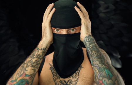 portrait of a guy in a balaclava with a bare torso in tattoos holding his headの写真素材