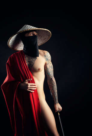 a naked slender man samurai in a red cape and an Asian hat with a tattooed katanaの写真素材