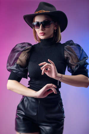 fashionable stylish woman in black clothes and hat poses with neon backgroundの写真素材