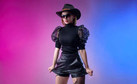 fashionable stylish woman in black clothes and hat poses with neon backgroundの写真素材