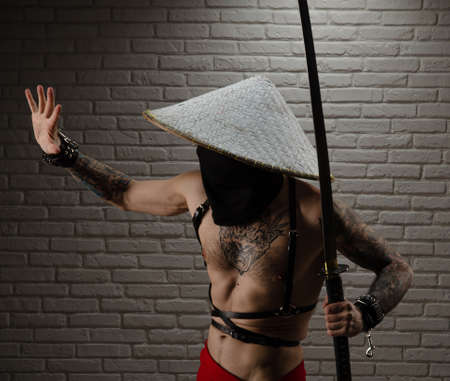 samurai man in a red cape and an Asian triangular hat with tattoos and a katana sword in his hands against the background of a brick wallの写真素材