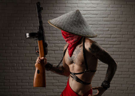 man in an Asian triangular hat with tattoos and a PPSh-41 airsoft gun and against the background of a brick wallの写真素材