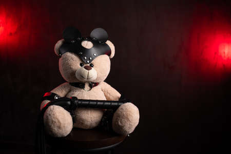 a toy teddy bear with a whip, dressed in leather belts and a mask, an accessory for BDSM games on a dark red texture backgroundの写真素材