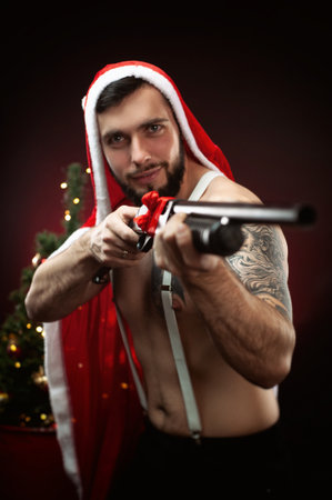 a strong guy in a red Santa Claus cape at the Christmas tree with a shotgun in his handsの写真素材