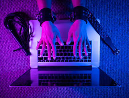 female sexy hands in bdsm handcuffs with laptop in neon light on dark background