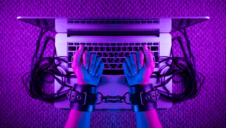 female sexy hands in bdsm handcuffs with laptop in neon light on dark background