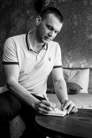 A young man makes a note in a notebook on a wooden table. A man is sitting on a bed in a loft-style room. A black-and-white photo of a businessman against the background of a stylish interior.の写真素材