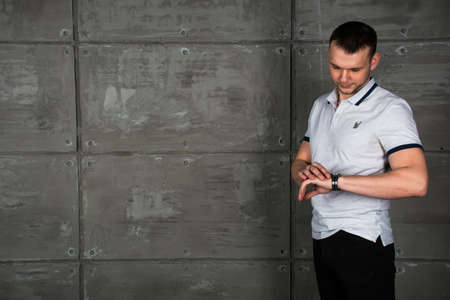 The young man looks at his watch. A man stands in a loft-style room on a gray background. Stylish interior.の写真素材