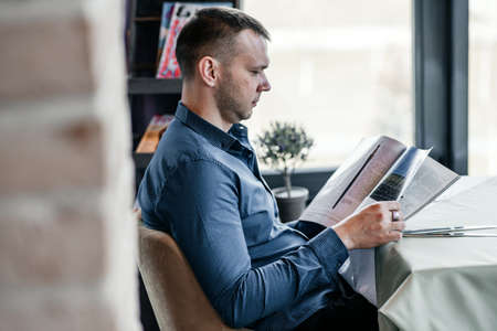 A young man in a blue shirt is reading a magazine in a cafe. A businessman is waiting for breakfast in a cozy cafe with a pleasant interior.の写真素材