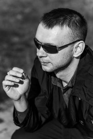 A young man in dark glasses looks into the distance. A businessman in a stylish raincoat is sitting on a bench and intends to take off his sunglasses. Black and white photo.の写真素材
