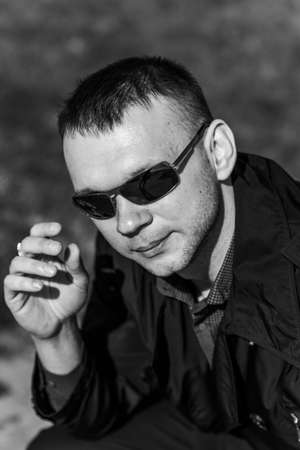 A young man in dark glasses is resting on a bench. A businessman in a stylish raincoat takes off his sunglasses. Black and white photo.の写真素材