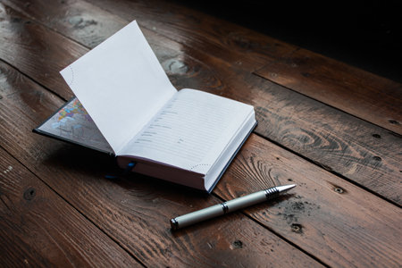 A black notebook and silver pen on a brown lacquered Desk. The first page is open.の写真素材