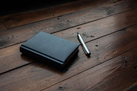 A closed notebook and a silver pen on a lacquered wooden table.の写真素材