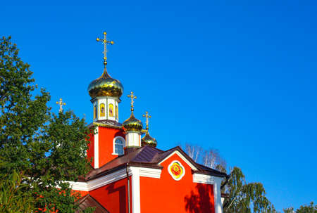 Red, Orthodox church in the park, against the blue sky. A religious building with golden domes and crosses stands around green trees.の写真素材
