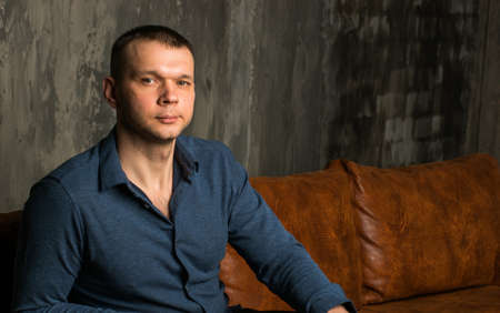 A young man in a blue shirt is sitting on a vintage sofa in a dark room, against a brick wall.の写真素材