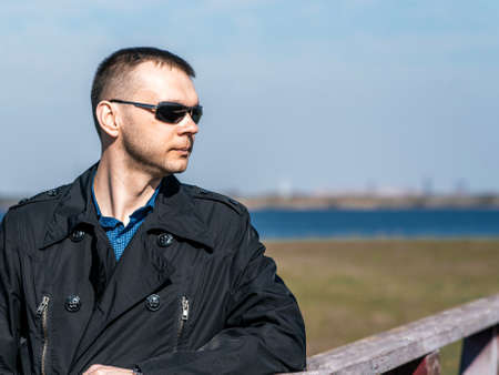 A young man in a black coat and sunglasses stands on the bank of the river and looks into the distance. A young, handsome businessman on an old, wooden bridgeの写真素材